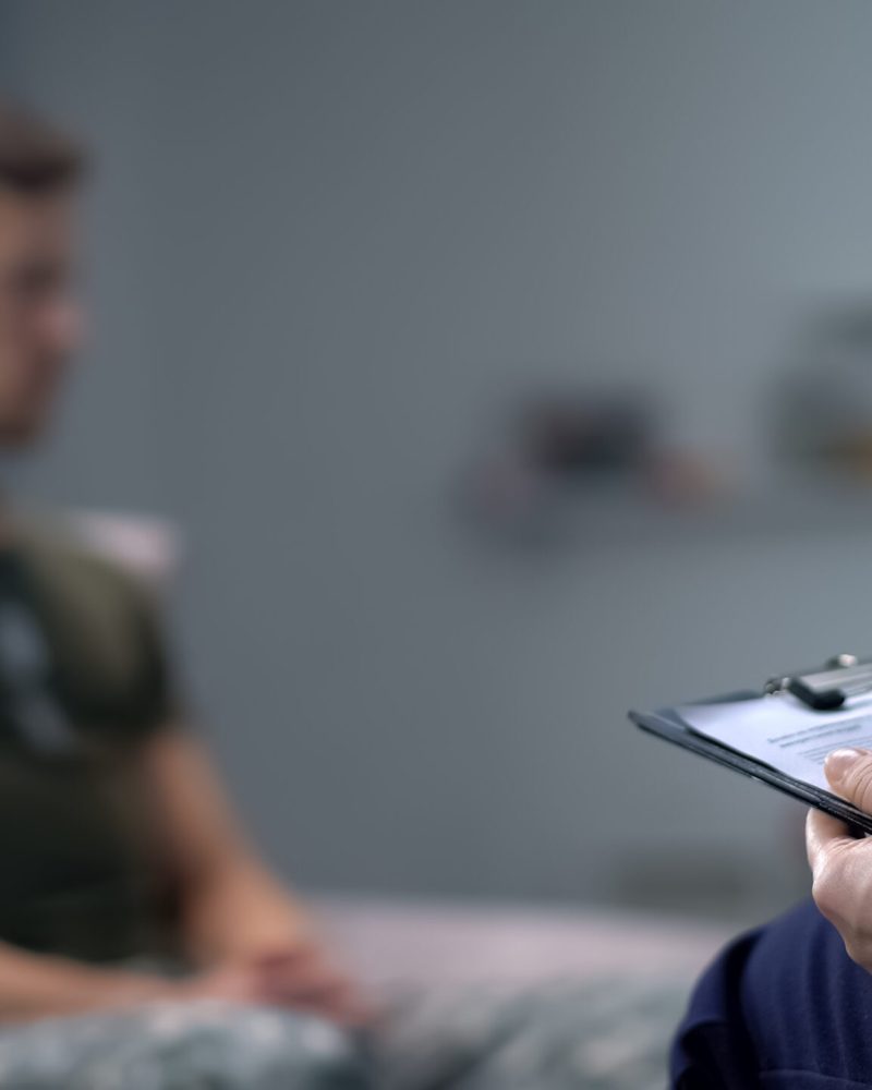 Psychologist,Making,Notes,During,Therapy,Session,With,Sad,Male,Soldier,